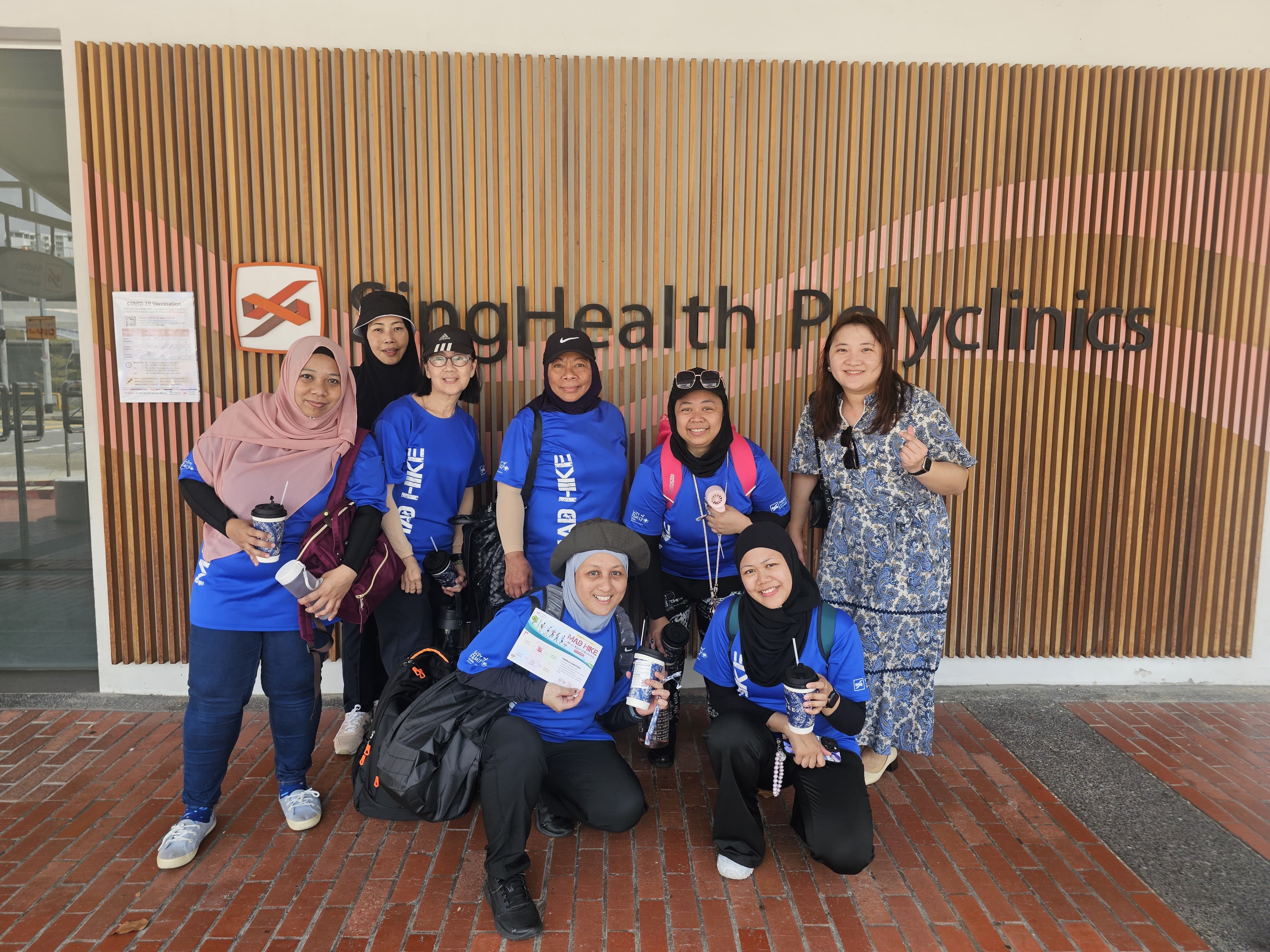 A group of MAD Hike participants in blue event T-shirts posing outside a SingHealth Polyclinics entrance.